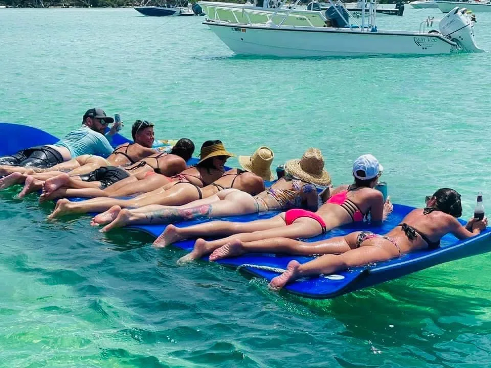 Group lounging on a floating mat over clear turquoise water near anchored boats, promoting Key West boat rentals, sandbar charters, tiki boat charters.