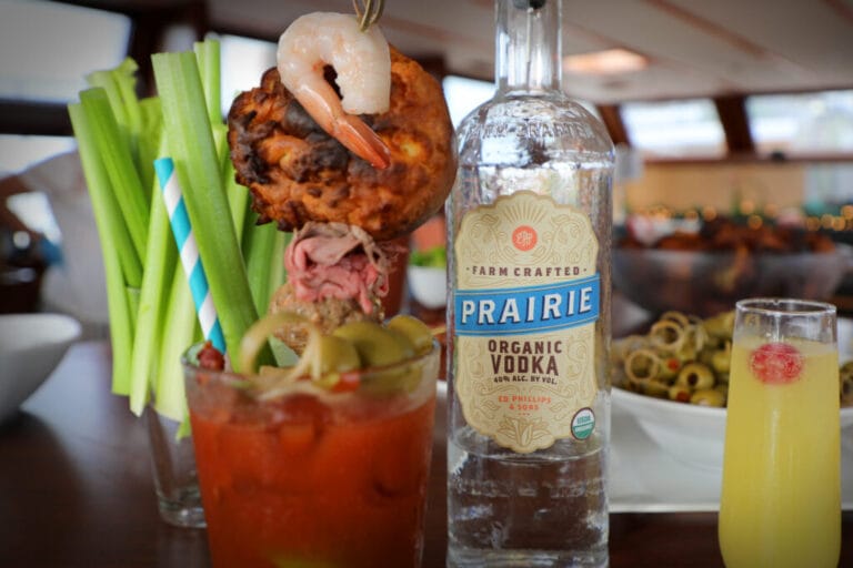 A transparent glass bottle of Prairie Organic Vodka displayed next to a fresh glass of orange juice on a wooden table, possibly suggesting a cocktail mix.